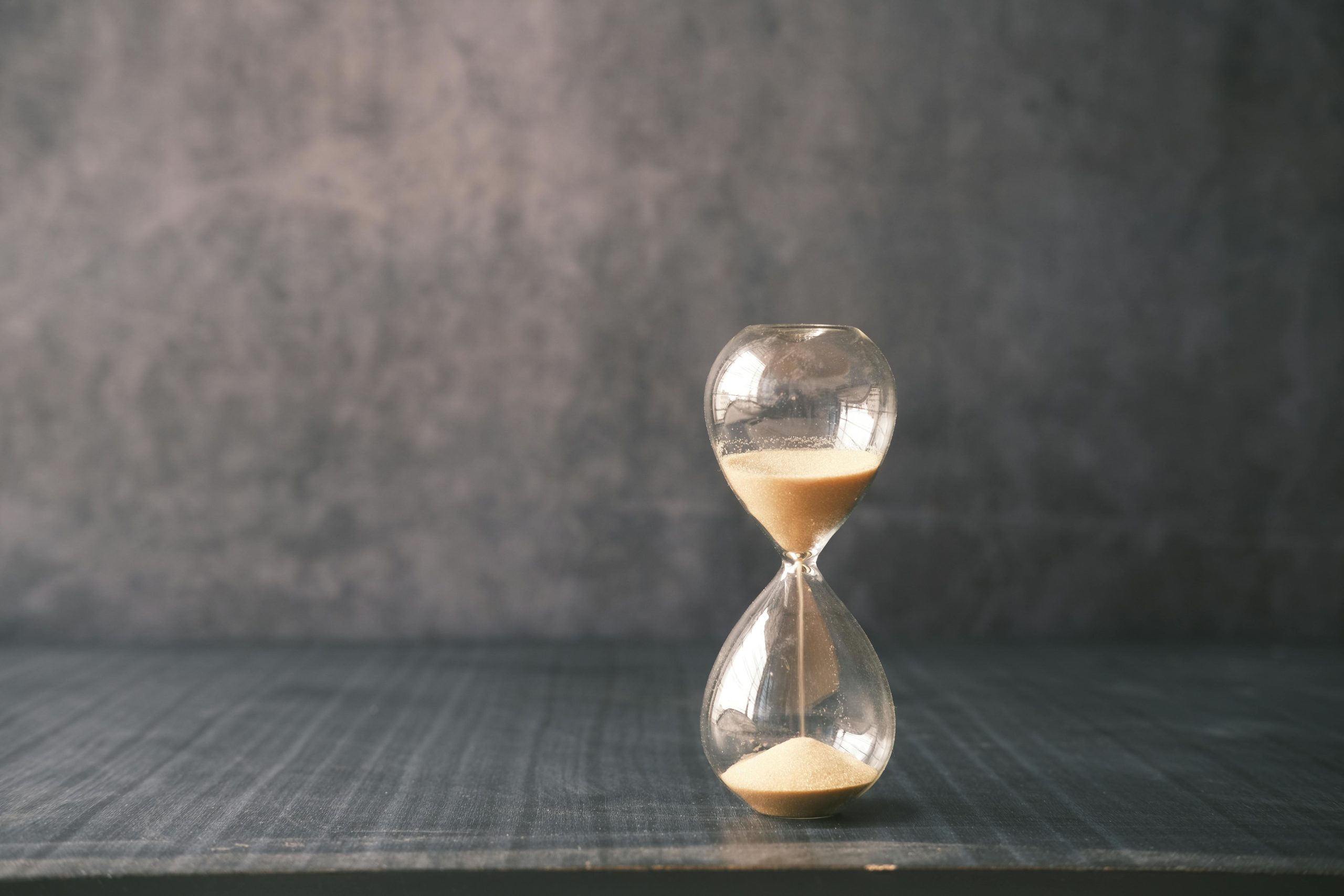 Time Running Out on Legacy Infrastructure A glass hourglass with sand flowing through it, resting on a dark wooden desk against a moody, textured gray background, symbolizing time running out on vulnerable legacy systems. - Destination Certification