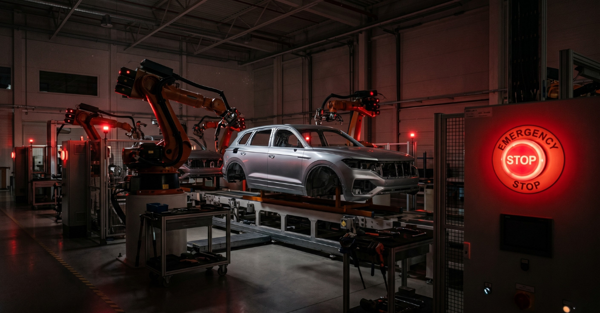 A glowing red emergency stop button in the foreground of a darkened, inactive high-end automotive assembly line, symbolizing production downtime. - Destination Certification