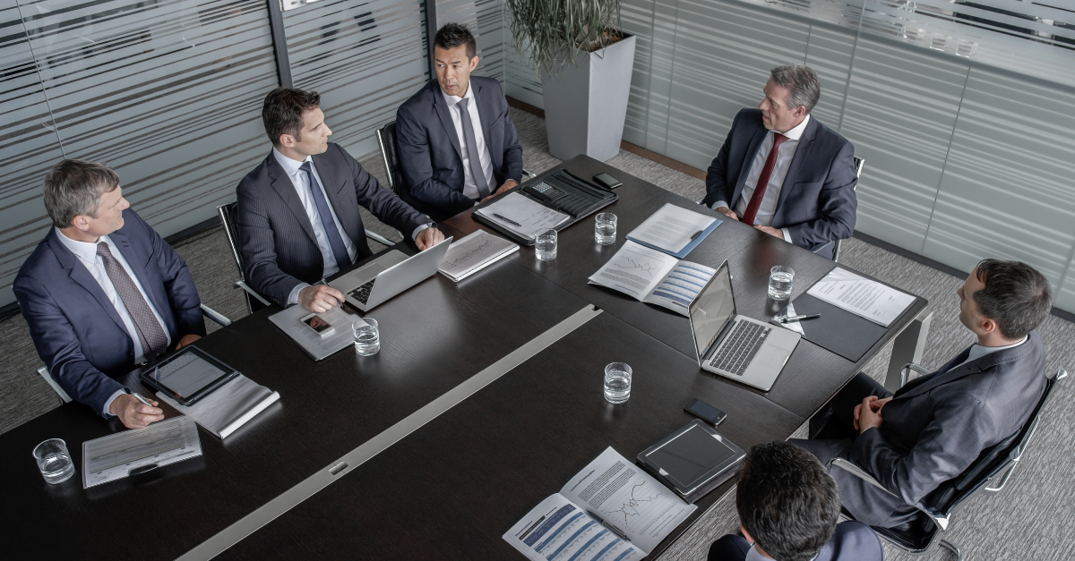 A high-angle photograph of six businessmen in suits seated at a dark wood conference table, engaged in a discussion in a modern office with glass walls. - Destination Certification
