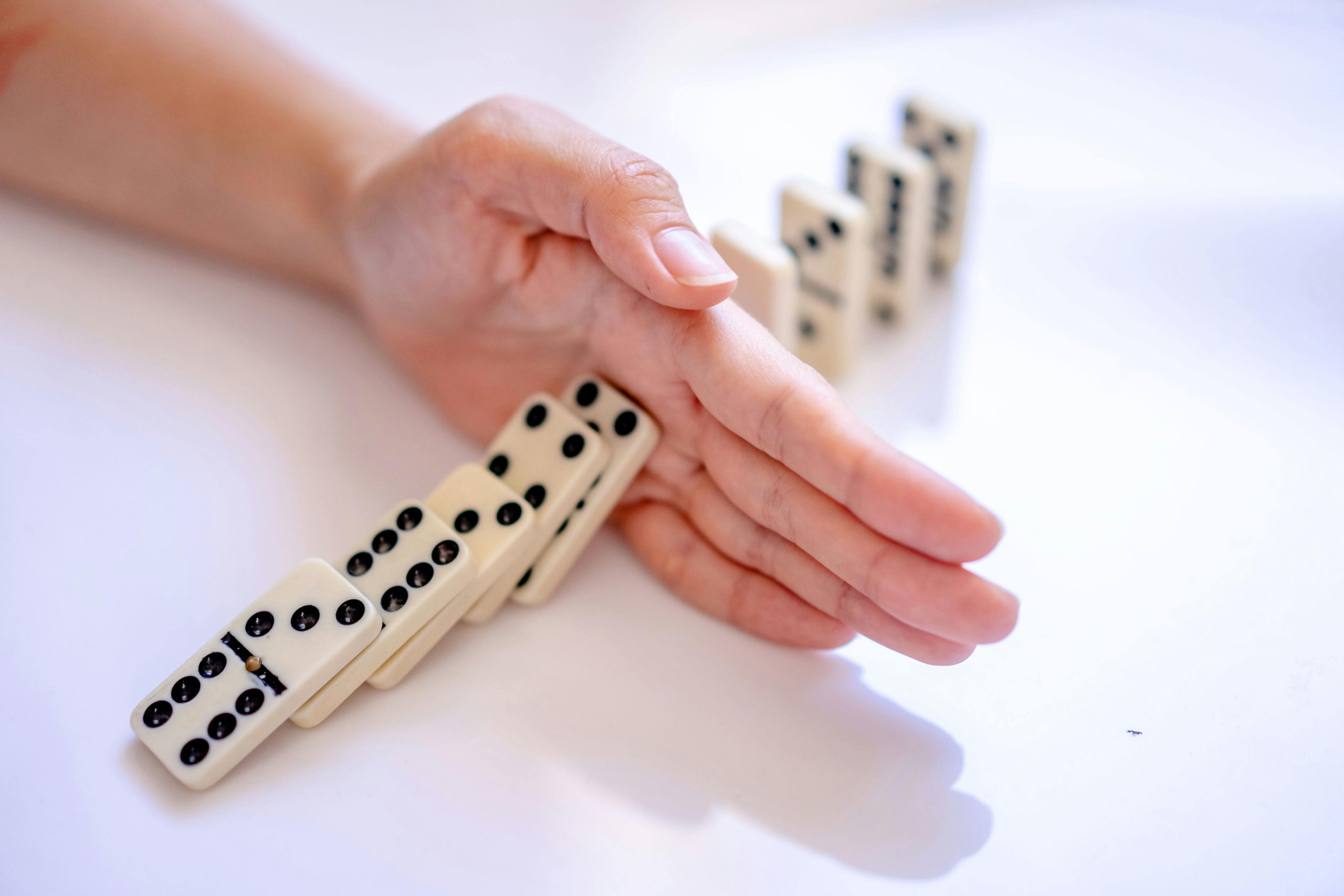 A close-up of a human hand placed vertically to intervene and stop a chain reaction of falling dominoes. - Destination Certification
