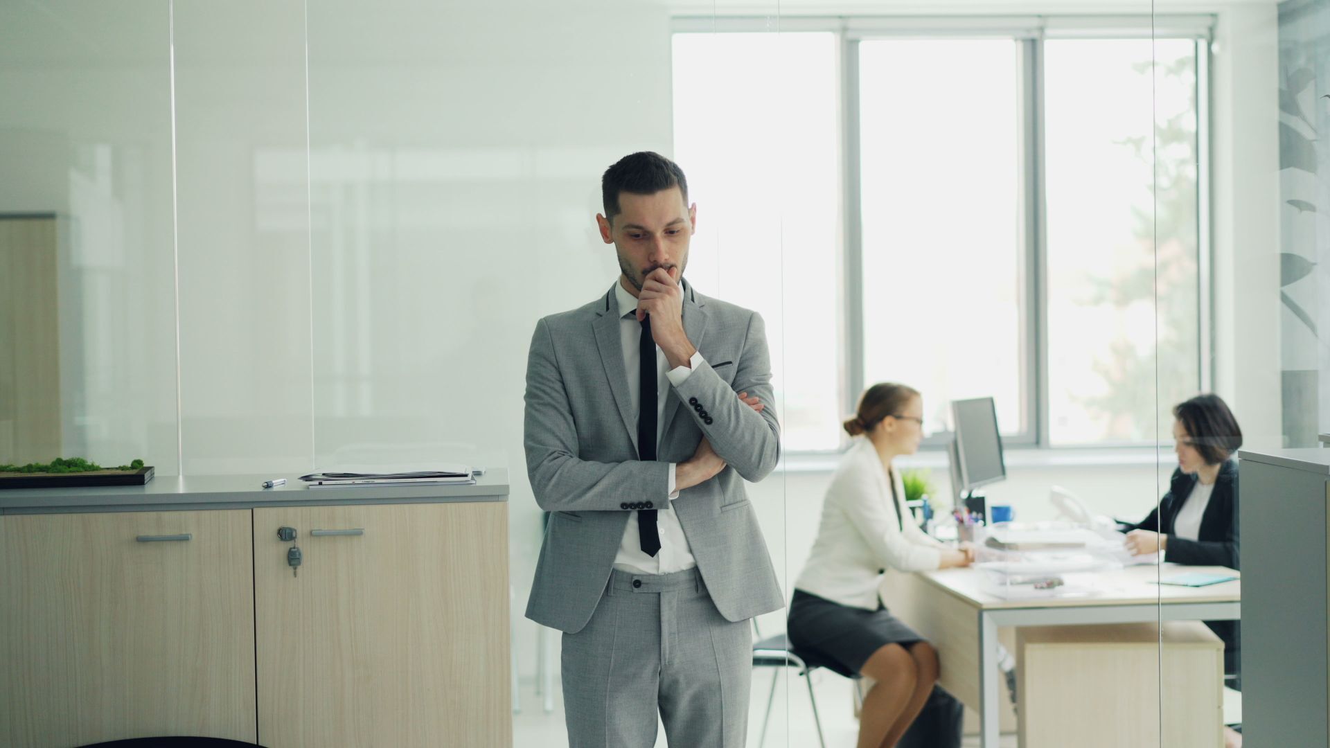 A man in a grey suit stands thoughtfully with his hand on his chin behind a glass partition in a modern office, while two women work at their desks in the background.